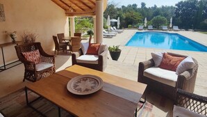 A covered outdoor seating area with wicker furniture, a wooden table, and a view of a swimming pool.