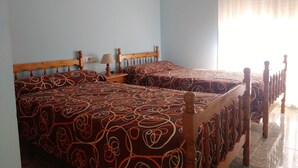 Una habitación con dos camas de madera, ropa de cama estampada, una mesita de noche con lámpara y una ventana con cortinas traslúcidas.