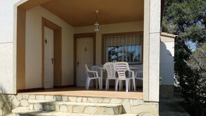 Un espacio al aire libre techado con una escalera de piedra que lleva a una puerta, una mesa con sillas de plástico y una ventana con barrotes.
