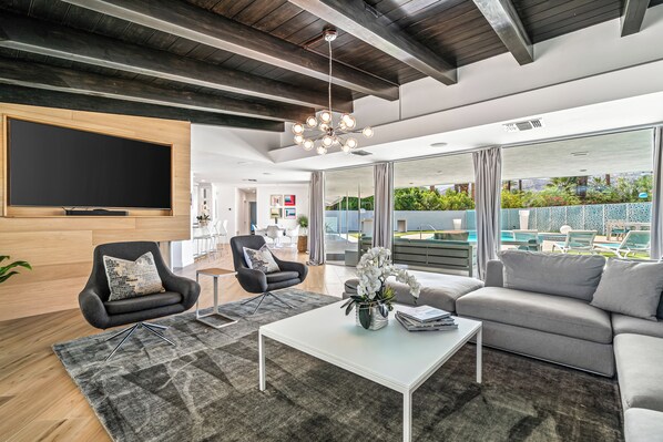 A modern living room with a large flat-screen TV, a sofa, a coffee table, and a dining area in the background.