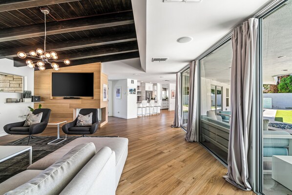 A modern living room with a large TV, wooden flooring, and a dining area with white furniture. - Lucy & Desi's Desert Escape | A Louisa Davis Property (Rancho Mirage)