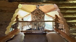 Interior rústico de una cabaña con chimenea de piedra, techo de madera y un balcón con barandilla.