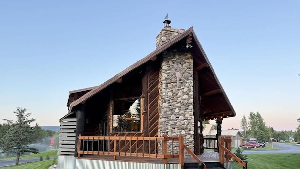 Una cabaña de troncos con chimenea de piedra, terraza de madera y una escalera que lleva a la entrada.