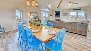 A dining area with a wooden table and blue chairs, a kitchen with a grey sofa, and a view of the city through large windows. - Oceanview Dog Friendly Home with Private Pool, Hot Tub, Game Room, and Wet Bar (Buxton)