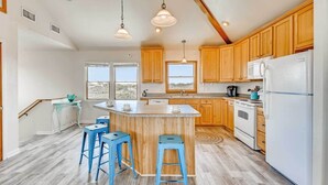 A kitchen with wooden cabinets, a white refrigerator, a central island with stools, and a view through the windows. - Oceanview Dog Friendly Home with Private Pool, Hot Tub, Game Room, and Wet Bar (Buxton)