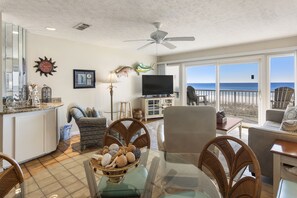 A spacious living room with a large glass table, wicker chairs, a television, and a view of the ocean.