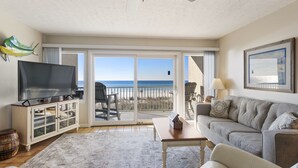 A living room with a television, a sofa, a coffee table, and a view of the beach through large sliding glass doors.