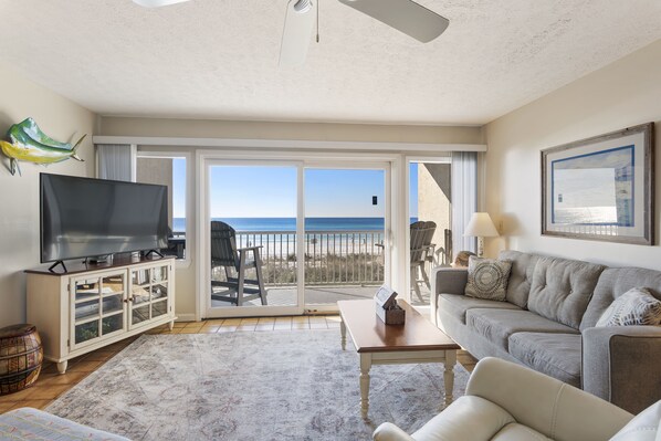 A living room with a television, a sofa, a coffee table, and a view of the beach through large sliding glass doors.