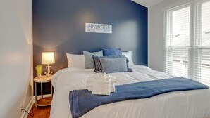 Ein ordentlich bezogenes Bett mit einer blauen Decke, ein Nachttisch mit Lampe und einer kleinen Pflanze, sowie ein Schild an der Wand mit der Aufschrift 'ADVENTURE'.