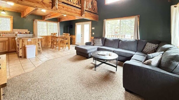 A spacious living area with a high ceiling, wooden beams, a large sofa, a coffee table, and a kitchen area with wooden cabinets and a dining table. - 24 MI to YNP | ATV Trails | Near Big Springs (Island Park)
