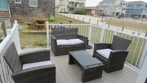 Eine Terrasse mit Korbmöbeln und einem Holztisch, mit Blick auf eine Rasenfläche und benachbarte Häuser.