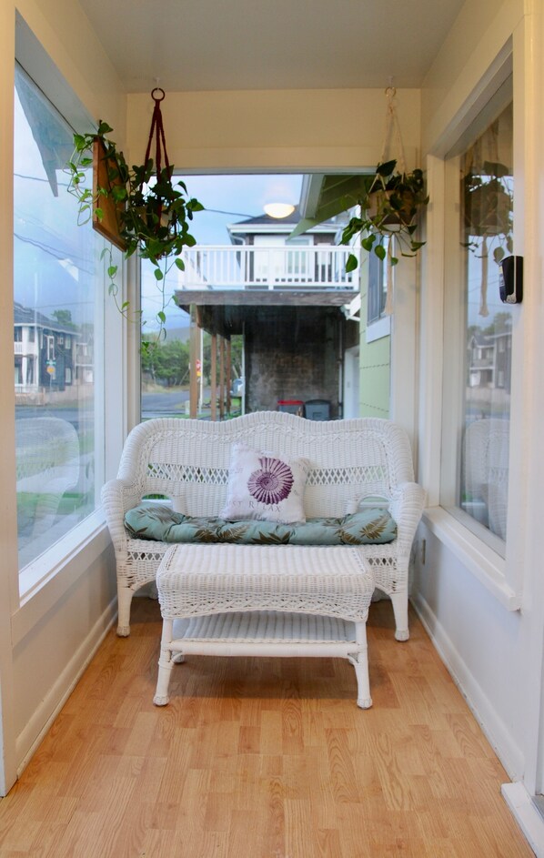 Un banco blanco de mimbre con cojín y plantas colgantes en una habitación con ventanas amplias.