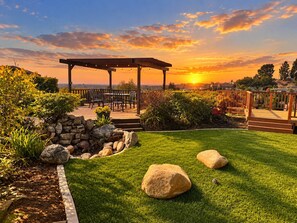 A garden patio with a pergola, seating, and a view of the sunset.