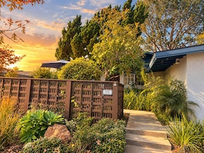 A gated entrance with a pathway leading to a building, surrounded by various plants and trees.