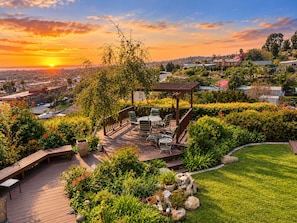 A deck with outdoor furniture, surrounded by lush greenery and a view of a cityscape at sunset. - Ocean & Bay Sunset Views from Family Oasis, Hottub (San Diego)