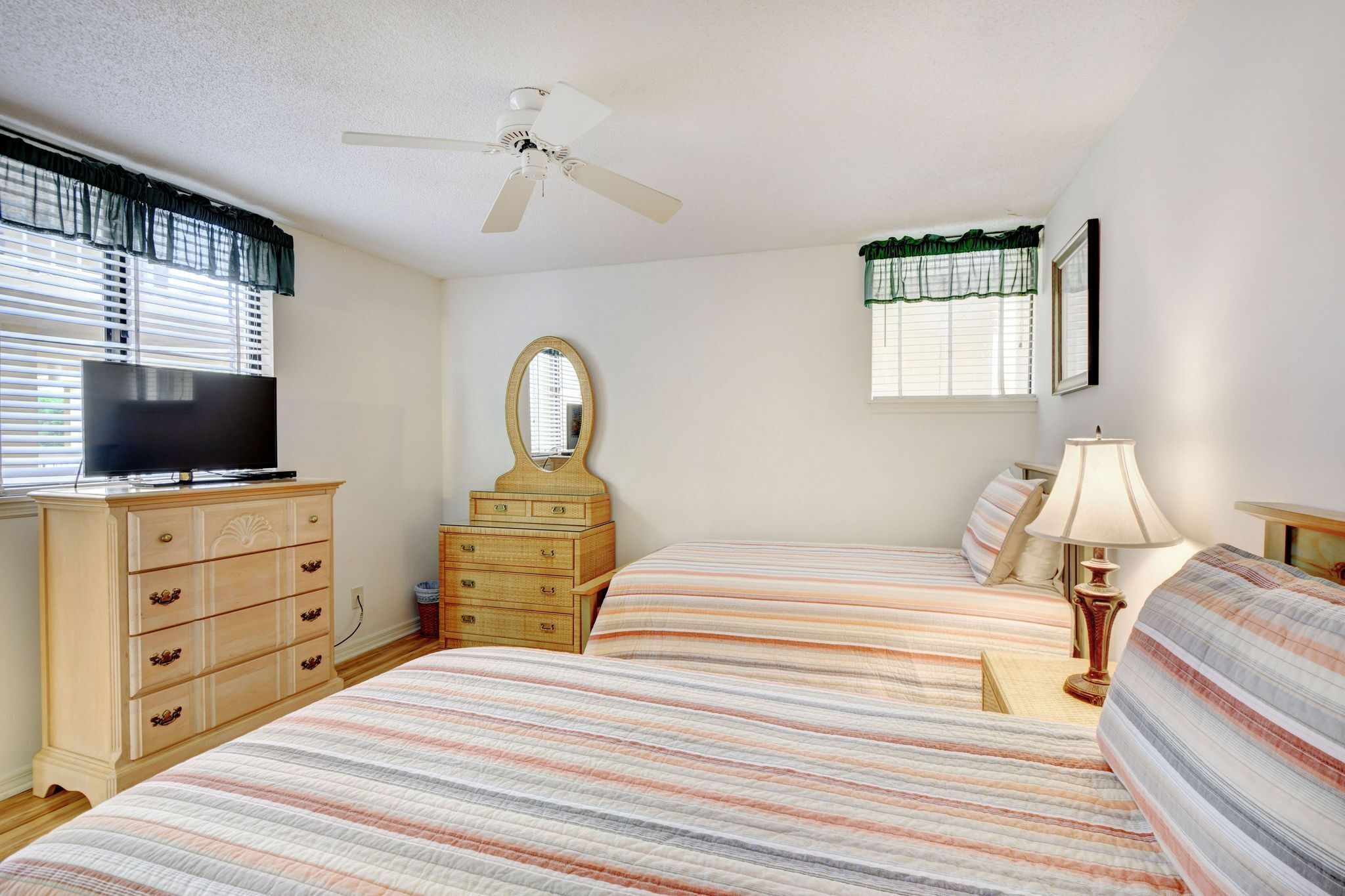 A bedroom with two beds, a dresser, a television, a ceiling fan, and a window with blinds.
