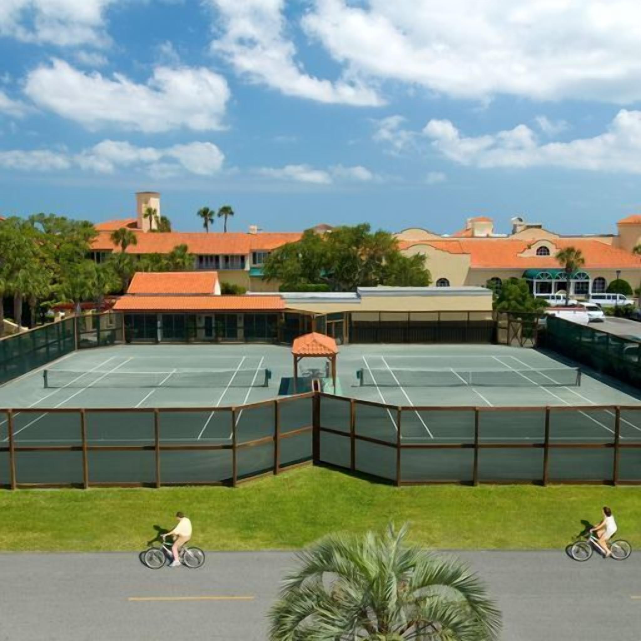 Un court de tennis entouré d’une clôture, un espace abrité avec des sièges, et deux cyclistes sur la route adjacente.