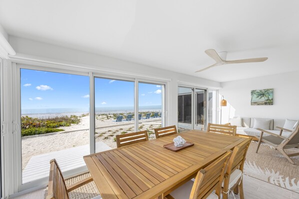 A spacious room with a large wooden table and chairs, a ceiling fan, and a view of the beach through large sliding glass doors.