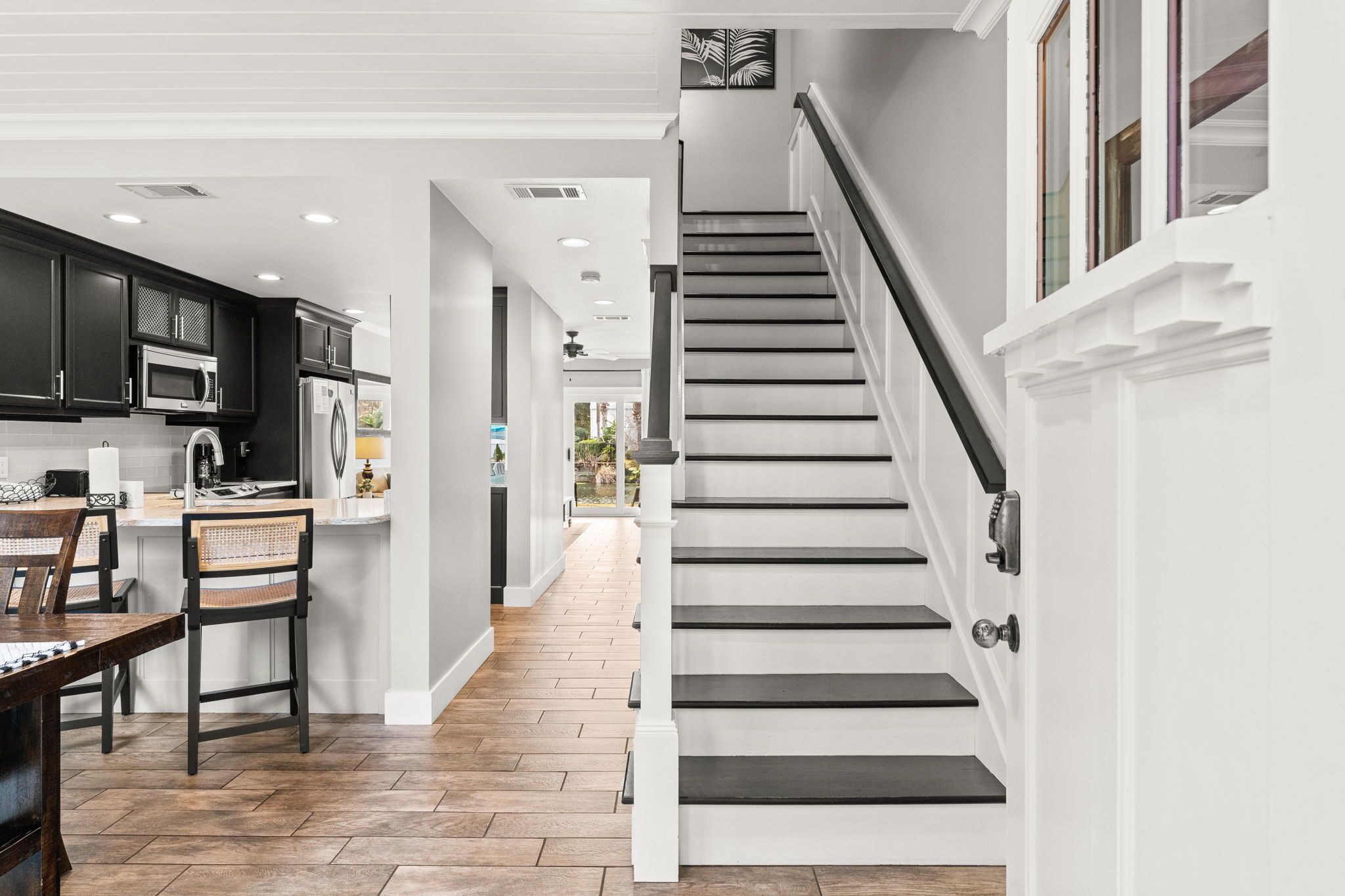 A modern kitchen with dark cabinets, a dining area with wicker chairs, and a staircase with white railings.