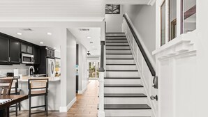 Una cocina moderna con armarios oscuros, un comedor con sillas de mimbre y una escalera con barandillas blancas.