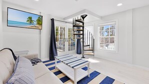 A modern living room with a sofa, a coffee table, a TV mounted on the wall, a staircase, and large windows. - Beautiful 5 Bedroom Captiva Pool Home Central to Stunning Beaches (Captiva)