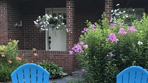Duas cadeiras Adirondack azuis em frente a uma casa de tijolos com porta e janela brancas, cercada por um jardim com várias flores.