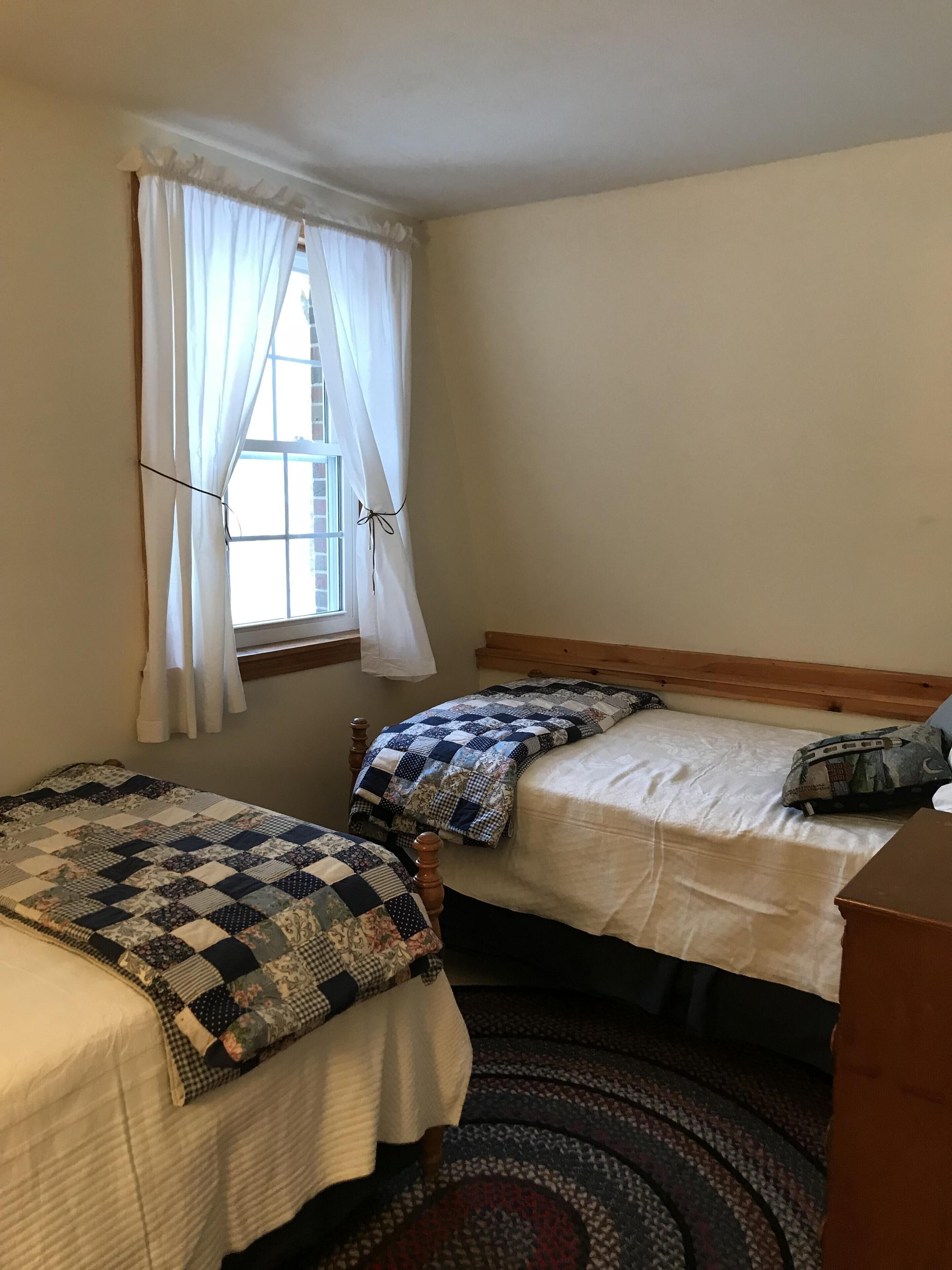 Une petite chambre agréable, dotée de deux lits simples, d’une fenêtre avec des rideaux et d’un tapis à motifs.