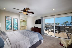 A bedroom with a large bed, a ceiling fan, a TV, and a balcony with a view of palm trees and a cityscape.
