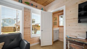 A cozy room with a black leather sofa, wooden walls, a white door, and a TV mounted on the wall. - Coastal Chic Cottage w/ View of Community Pool! (Port Aransas)