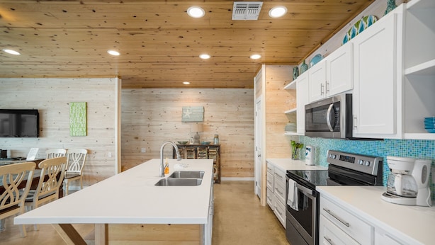 A modern kitchen with white cabinetry, a large island with a sink, and a microwave. There are wooden chairs and a dining table in the adjacent area. - Coastal Chic Cottage w/ View of Community Pool! (Port Aransas)