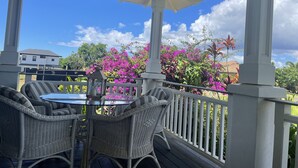 Un espacio al aire libre cubierto con muebles de mimbre y una mesa, rodeado por una baranda con vistas a una casa y plantas en flor.