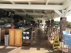A covered outdoor bar area with wooden furniture, a wet floor sign, and a view of the surrounding landscape. - HOME & GUEST HOUSE FOR PRICE OF 1 POIPU BEACH ESTATES 5 BR/ 5 BA WALK TO BEACH (Koloa)