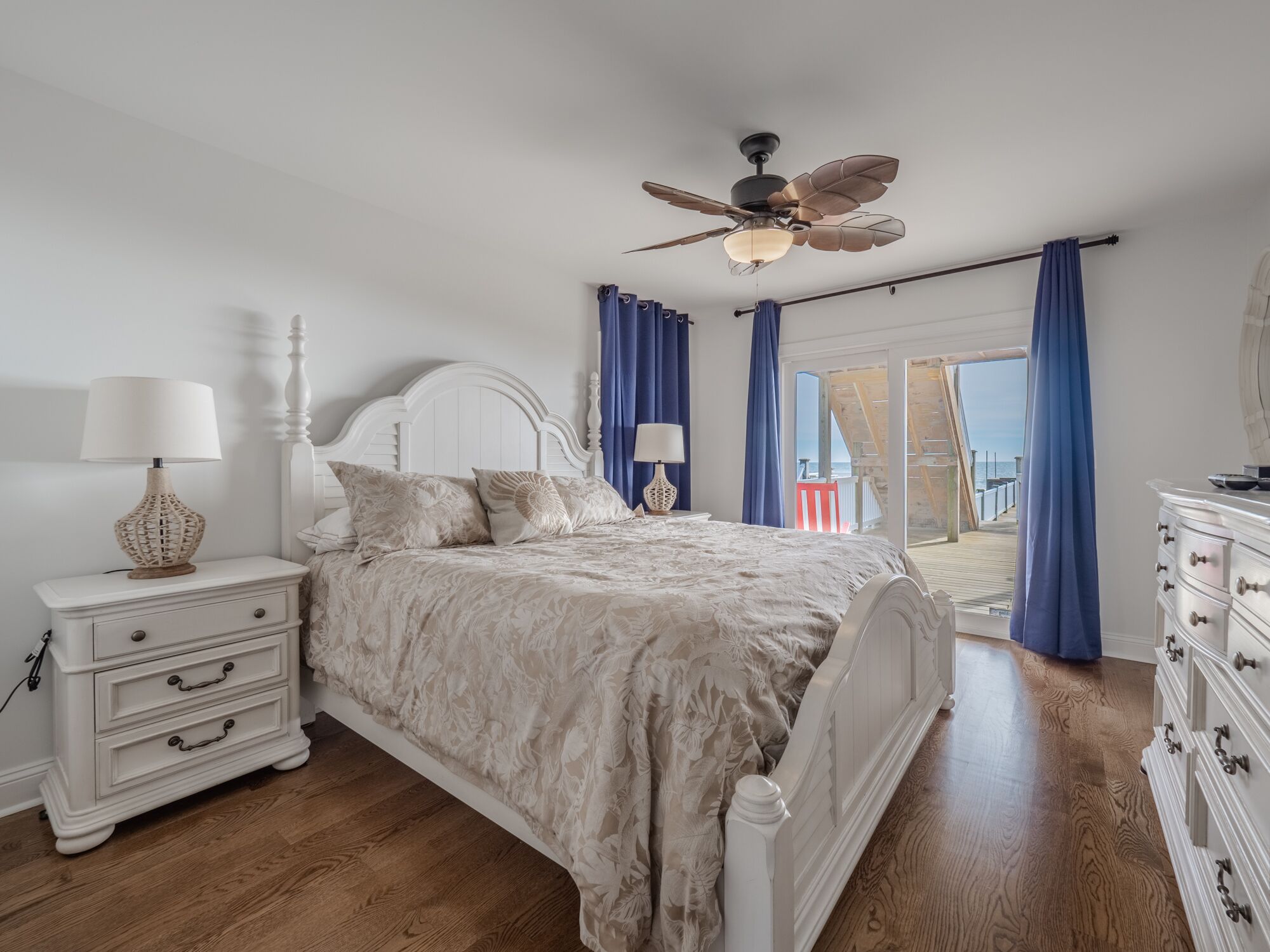 Un dormitorio con cama, mesitas de noche, cómoda, ventilador de techo y vista al mar desde un balcón.