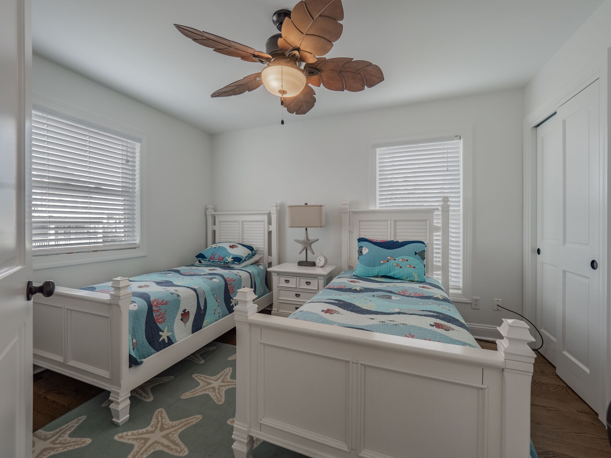 Une chambre avec deux lits, une moquette à motifs d’étoiles de mer, un ventilateur de plafond et des murs blancs.