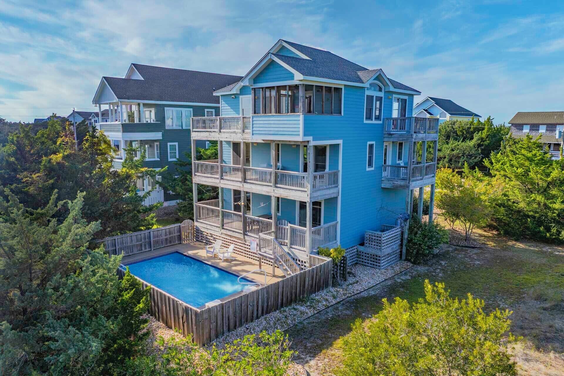 A multi-story house with a pool, surrounded by trees and other houses.