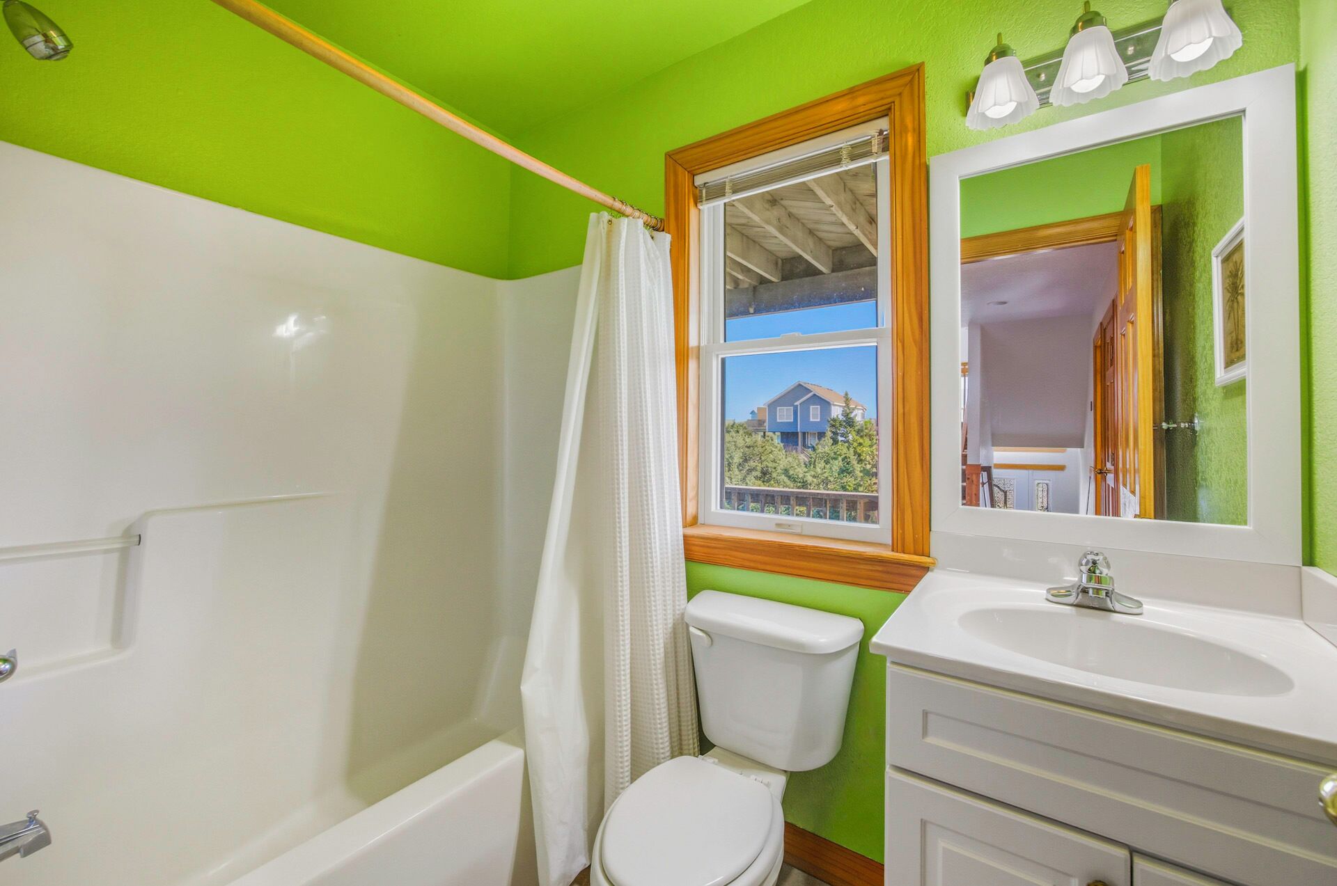 A bathroom with a green wall, a white toilet, a white sink, and a window with a view of a house.