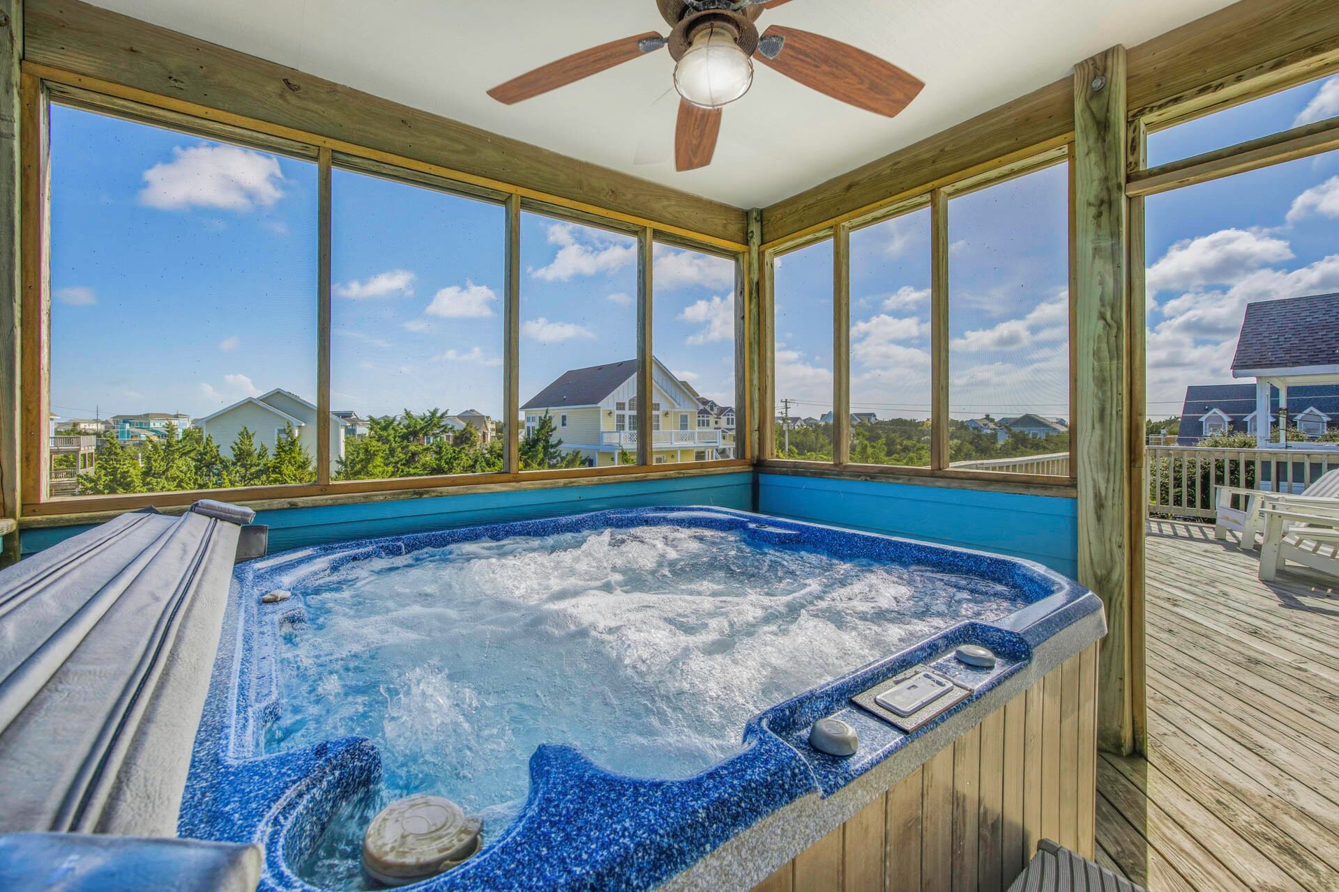 A hot tub with a wooden deck and large windows offering a view of houses and trees.