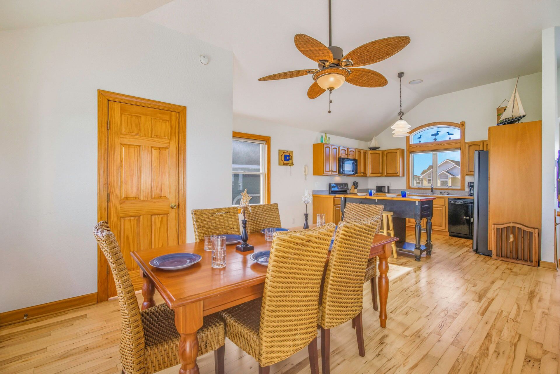 A dining area with a wooden table and wicker chairs, a kitchen with wooden cabinets and a black refrigerator, and a ceiling fan.