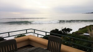 A seaside view from a balcony with a wooden table and chairs, overlooking the ocean with waves and a distant shoreline. - Panoramic Ocean View in a Beachfront Luxury Home (Santa Cruz)