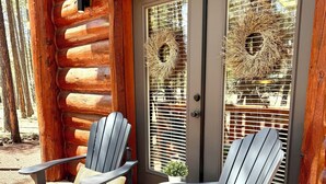 Dos sillas Adirondack sobre una terraza de madera frente a una cabaña de troncos con puerta y ventana.
