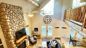 Amplia sala de estar con pared de piedra, chimenea, un sofá grande de cuero negro y una mesa de comedor de madera.