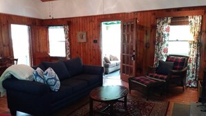 Un accogliente soggiorno con un divano blu scuro, un tavolino in legno e un tappeto decorato. Si apre una porta in legno verso un'altra stanza, e c'è una finestra con tende.