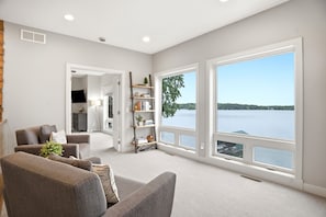 Een ruime woonkamer met een groot raam met uitzicht op een meer, een open haard en een boekenkast.