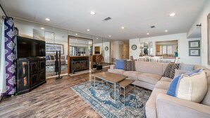 A spacious living room with a large sofa, a coffee table, and a TV. There is a kitchen area visible through a glass partition. - South Shore 2Bd, Lake View/Super Amenities/Mini-Golf/Tennis, Pickleball & More! (Hot Springs)