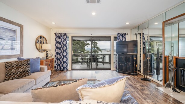 A spacious living room with a sofa, a coffee table, and a TV. There is a balcony with a table and chairs, and a fireplace with a TV. - South Shore 2Bd, Lake View/Super Amenities/Mini-Golf/Tennis, Pickleball & More! (Hot Springs)