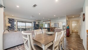 A dining area with a wooden table and white chairs, a sofa with patterned cushions, and a view through large windows. - South Shore 2Bd, Lake View/Super Amenities/Mini-Golf/Tennis, Pickleball & More! (Hot Springs)