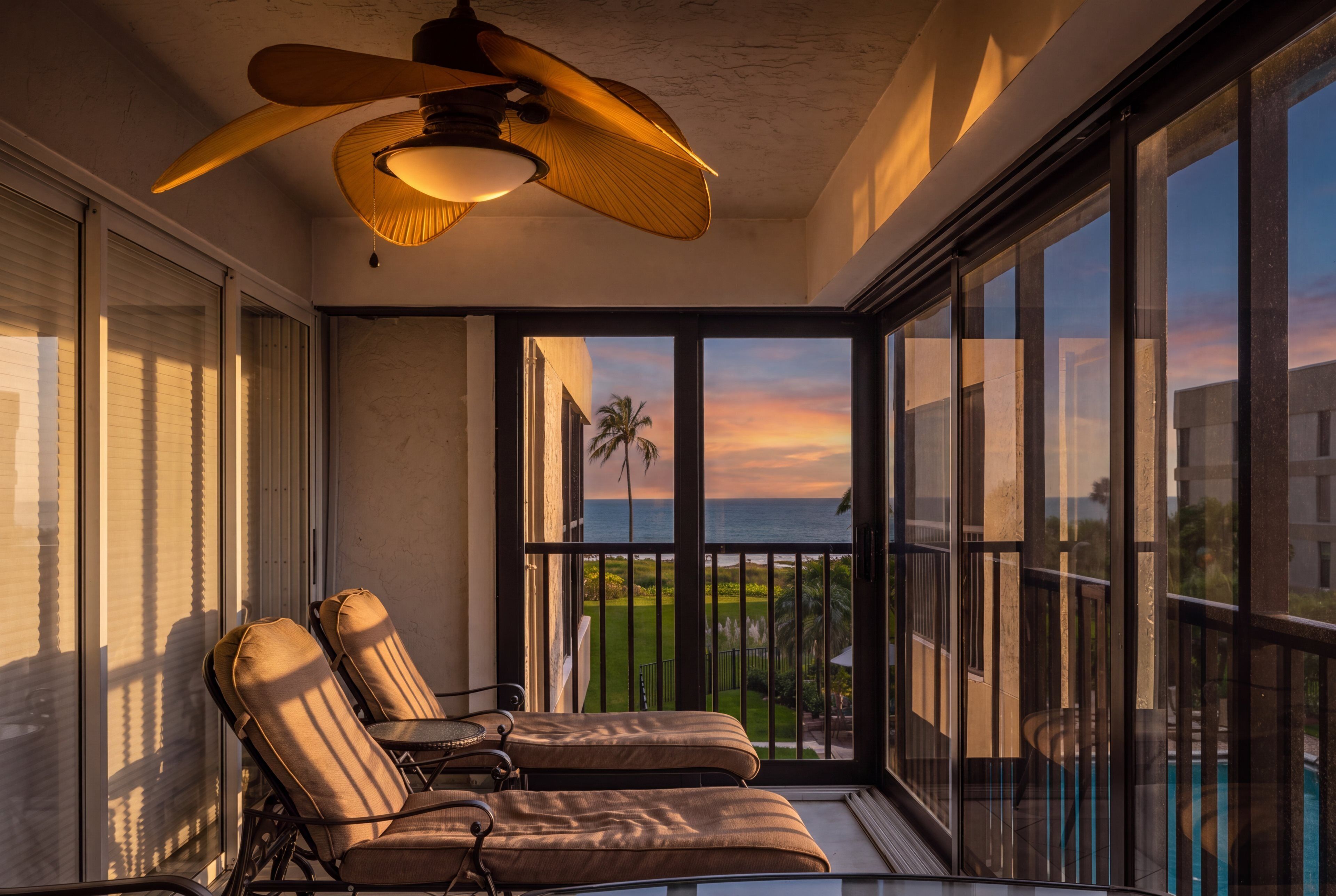 Ein Balkon mit zwei Loungestühlen, einem Deckenventilator und Blick auf den Ozean bei Sonnenuntergang.