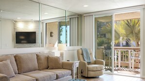 A modern living room with a beige sofa, a flat-screen TV mounted on a stone wall, a glass door leading to a balcony with a view of palm trees. - Stunning Ocean, Pool, and Garden views from Beautifully Appointed Unit! (Oceanside)