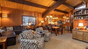 A cozy interior with a wooden ceiling, a living area with a sofa and ottoman, a dining table with chairs, and a kitchen area with wooden cabinets and a clock. - Grainger by Hauserman Rental Group (Tahoe City)