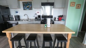 A kitchen with a wooden island, four black stools, stainless steel appliances, and decorative wall art. - LastMinuteSpecial/OceanFront/Pool/HotTub/XBOXoneS/ping pong table (Nags Head)
